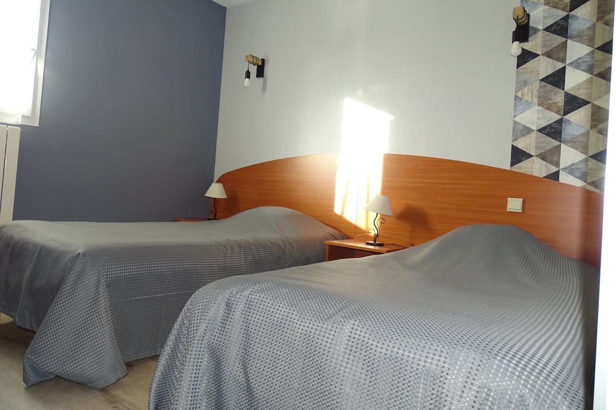 Habitación de hotel con dos camas, cabecera de madera, mesita de noche con lámpara y pared con estampado geométrico.