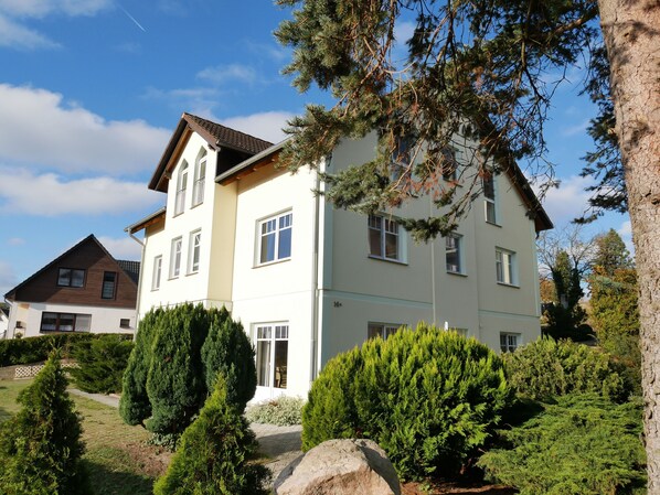 A two-story house with a sloped roof, multiple windows, and a well-maintained garden with various shrubs and a large rock.