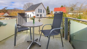 Een balkon met een ronde tafel en twee stoelen, een vaas met bloemen en een uitzicht op huizen op de achtergrond.