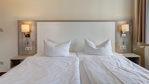 Een hotelkamer met twee bedden, elk met witte beddengoed en kussens, en wandlampen aan weerszijden.