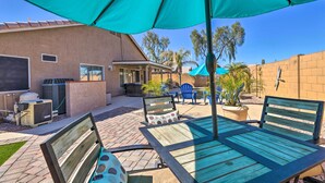 An outdoor patio area with a table and chairs under a large umbrella, surrounded by a brick wall and a covered seating area. - Outdoor Kitchen, Heated Pool: Gilbert Retreat! (Gilbert)