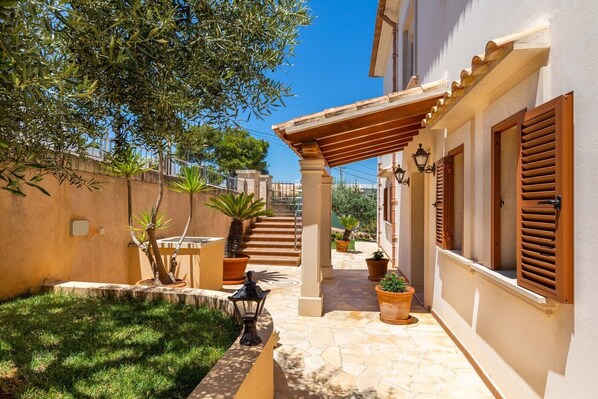 A courtyard with a stone floor, potted plants, a pergola, and a staircase leading to a building with wooden shutters.