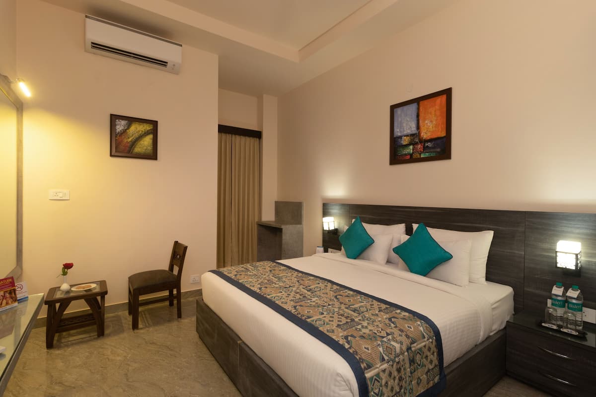 Una habitación de hotel con una cama grande, una silla, una mesita con un jarrón de flores y una mesita de noche con botellas de agua.