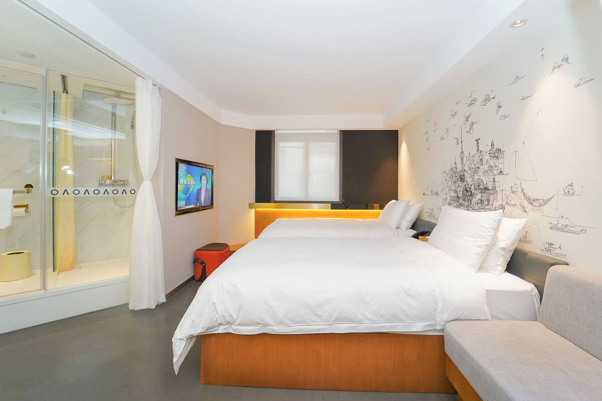 A modern hotel room with a large bed, a glass shower enclosure, a small table, and a wall with a cityscape mural.