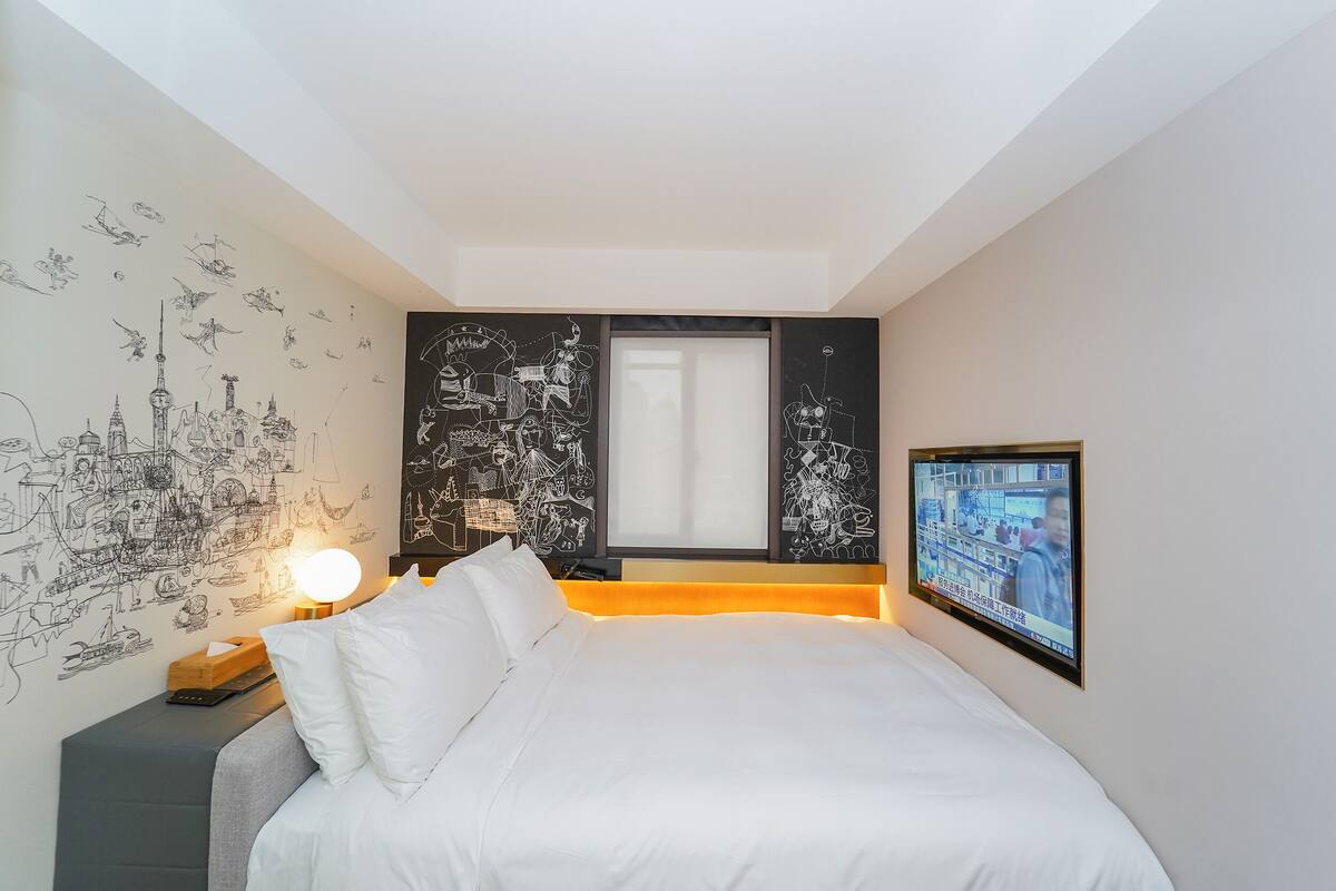 A hotel room with a bed, a bedside table with a lamp, a wall with cityscape drawings, and a TV on the wall.
