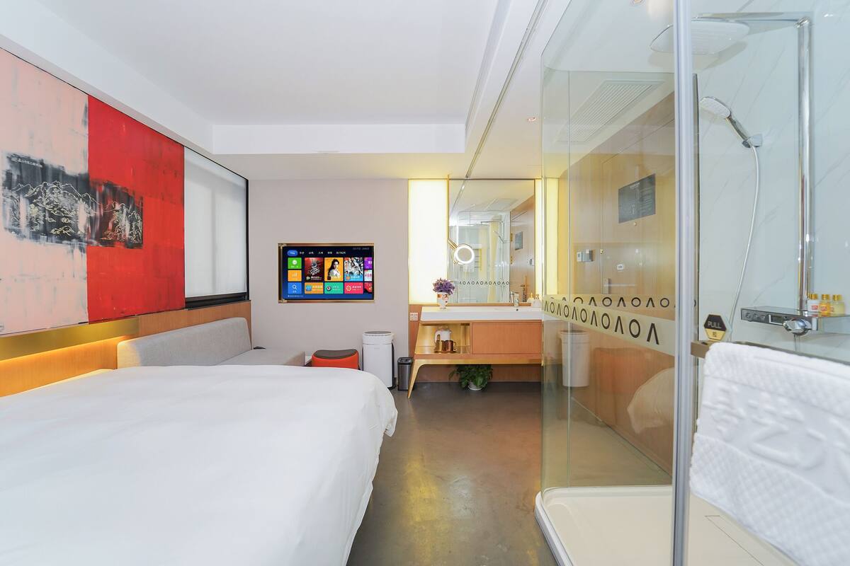 A modern hotel room with a large bed, a TV mounted on the wall, a bathroom with a glass shower enclosure, and a red abstract painting on the wall.