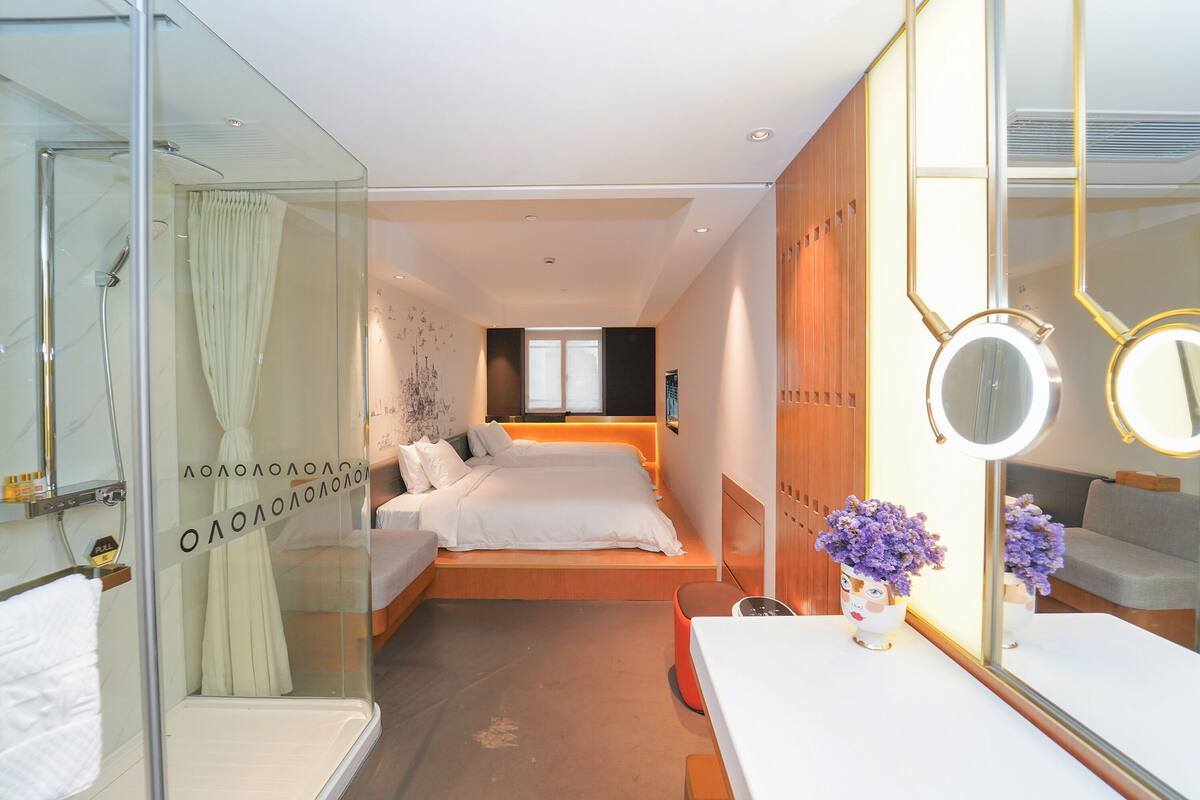 A modern hotel room with a glass shower enclosure, a bed with white bedding, a bedside table with a mirror, and a small orange stool.