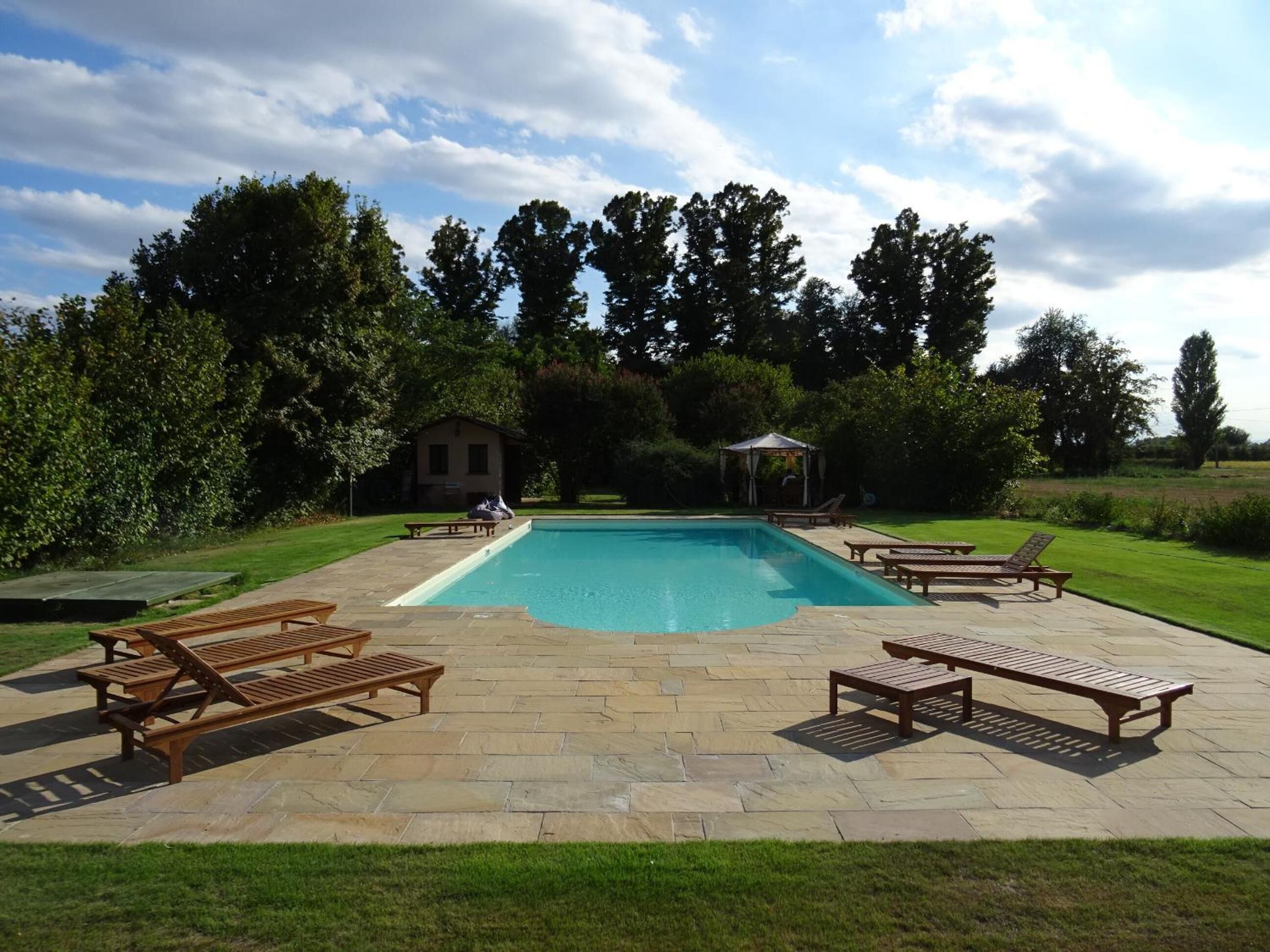 A rectangular swimming pool with a stone deck, surrounded by wooden loungers and a gazebo.