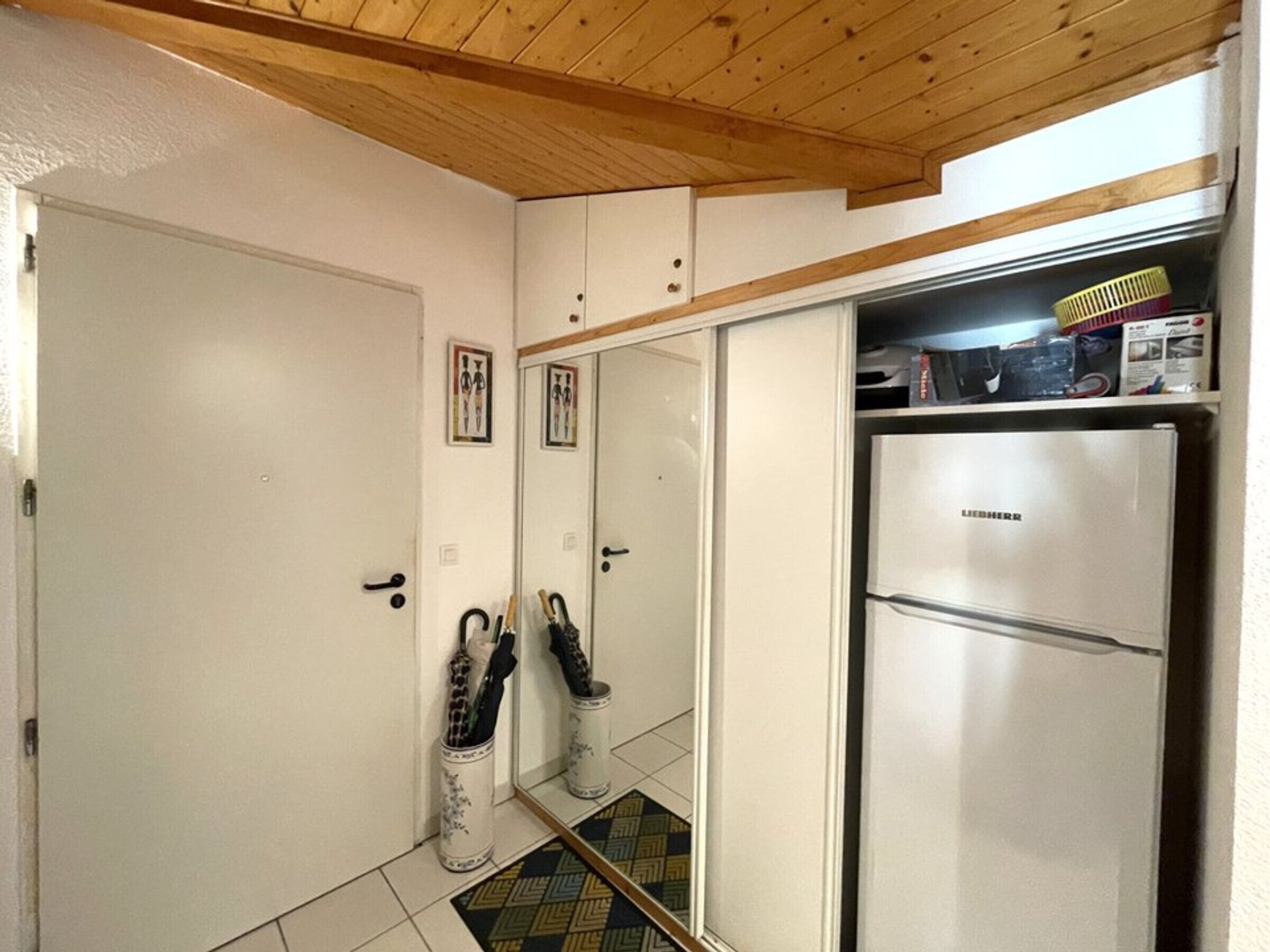 A compact kitchen with a stainless steel refrigerator, a wooden ceiling, and a tiled floor.