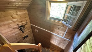 Interior de una cabaña de madera con escalera en espiral, cabeza de venado montada en la pared y una ventana con mosquitero.