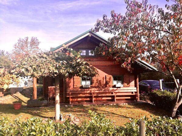 A traditional wooden house with a sloped roof, a front porch, and a garden area with potted plants and a tree.