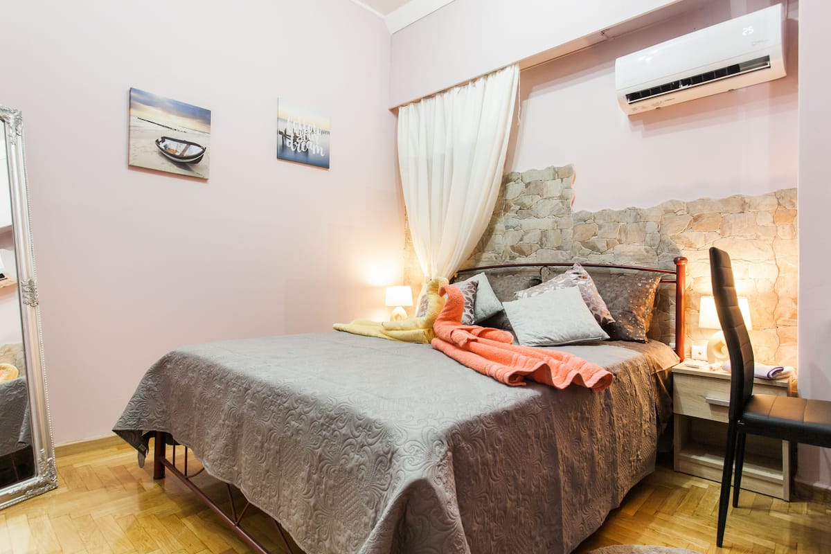 Une chambre à coucher avec une tête de lit en pierre, un lit recouvert d’une couverture grise, une table de chevet avec une lampe, une chaise et un miroir.