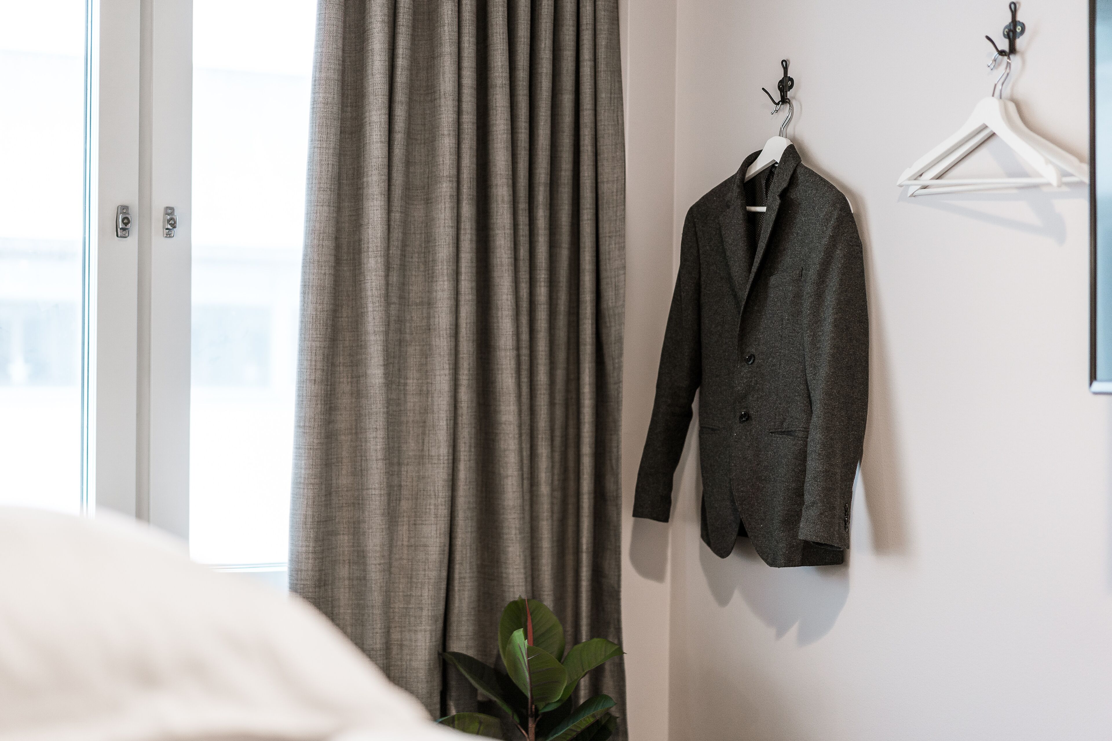 A room with a bed, a grey curtain, a grey coat hanging on a wall, and a plant.