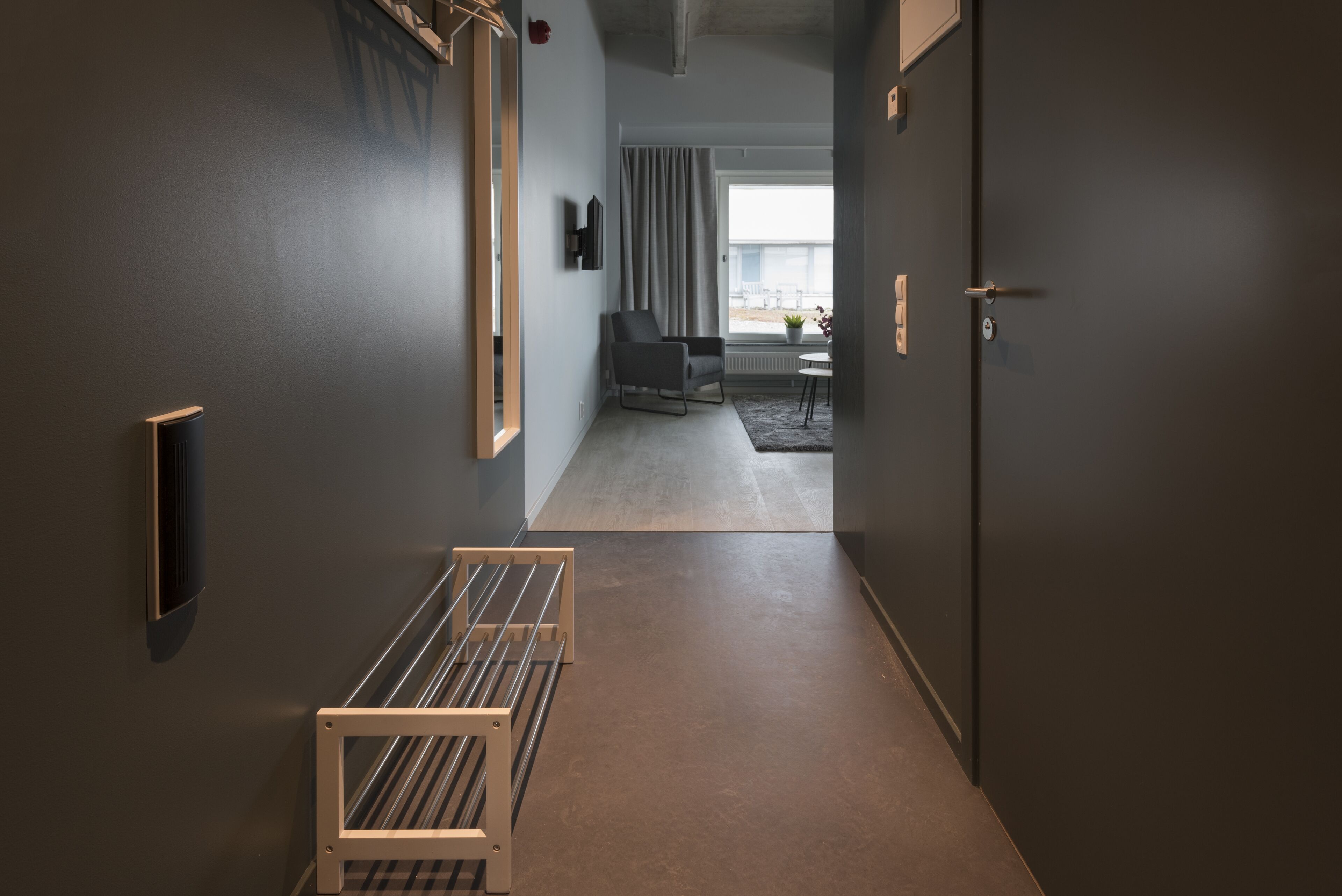 Un pasillo moderno de hotel con un banco, una silla y vistas a una habitación con un ventanal y cortinas.