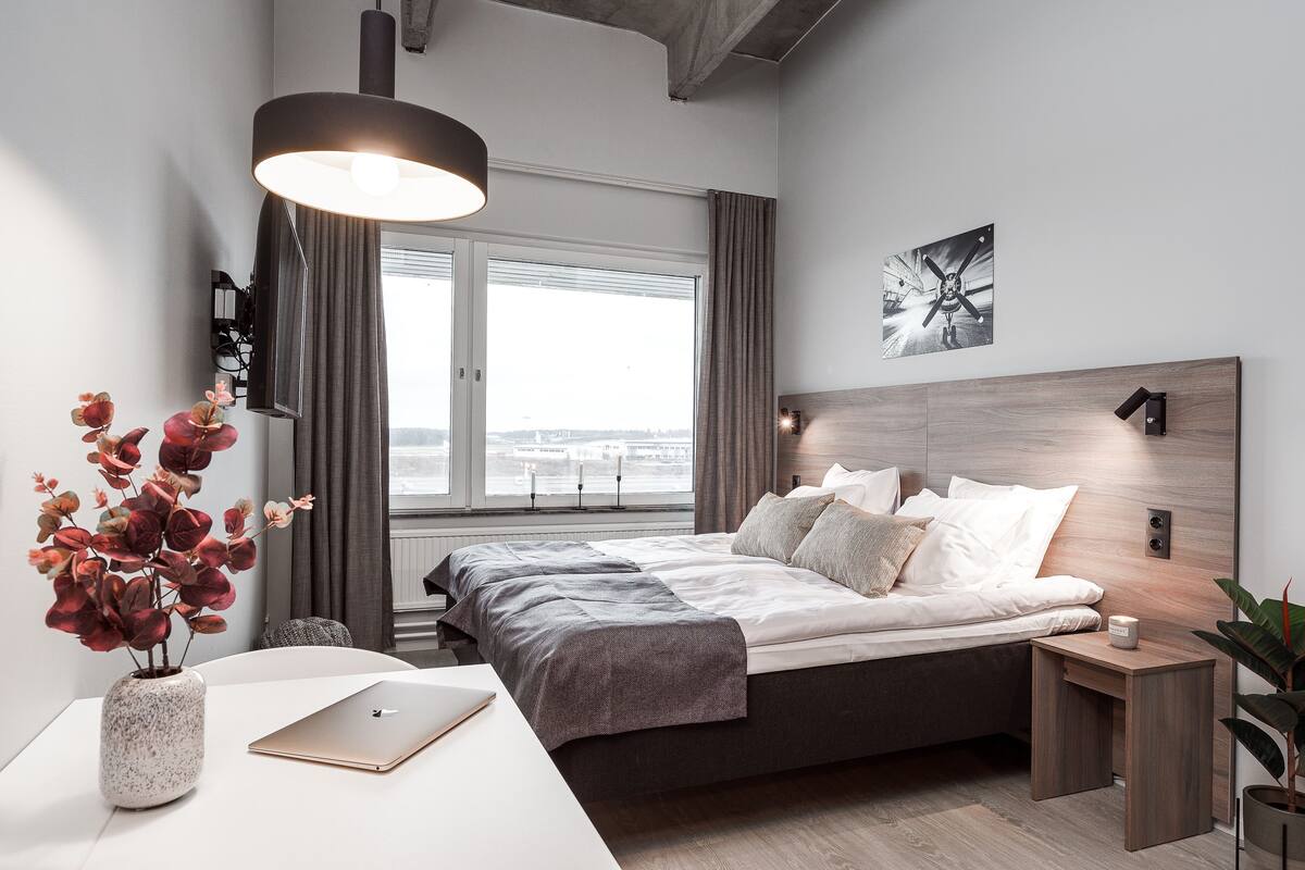 Ein modernes Schlafzimmer mit einem Bett, Nachttischen, einem Schreibtisch, einem Laptop, einer Vase mit Blumen, einem Fenster mit Vorhängen und Blick auf die Stadt.