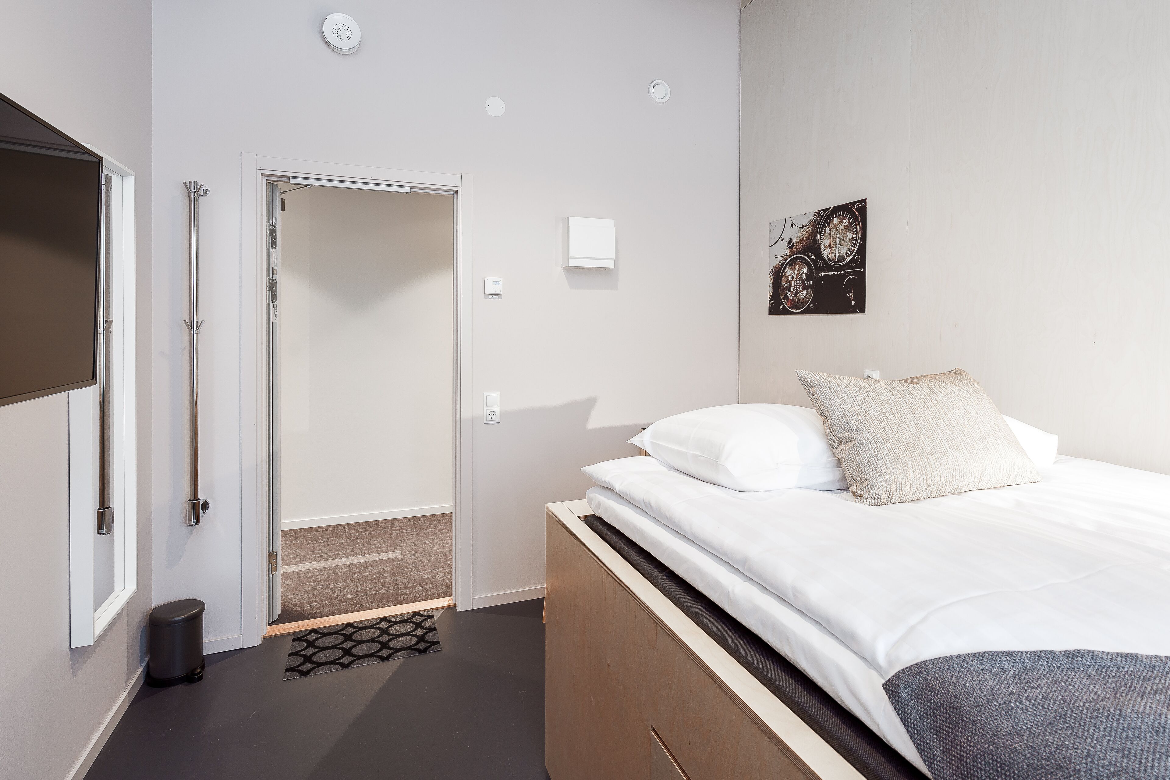 Una habitación de hotel moderna con una cama, un cesto de basura pequeño, una puerta y una lámpara fijada a la pared.