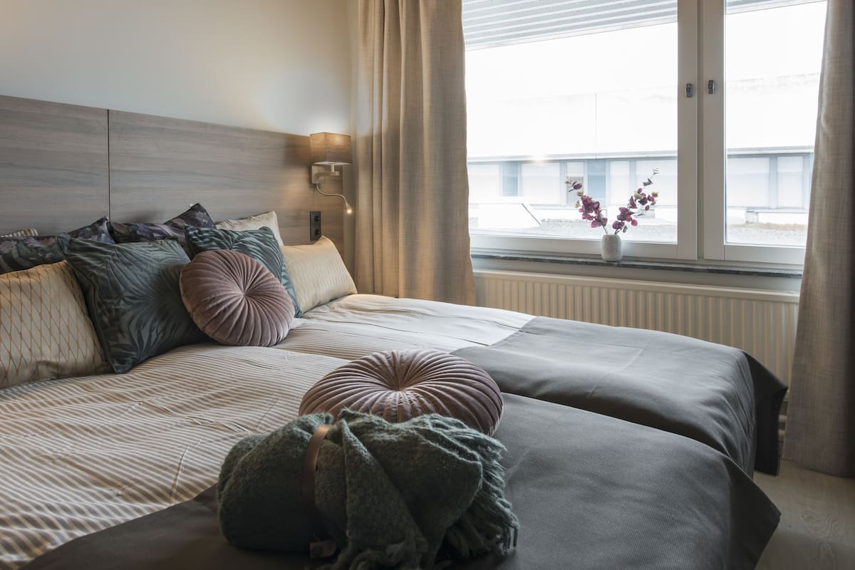 Ein ordentlich bezogenes Bett mit mehreren Kissen und einer Decke, eine Nachttischlampe und ein Fenster mit Blick auf ein Gebäude.