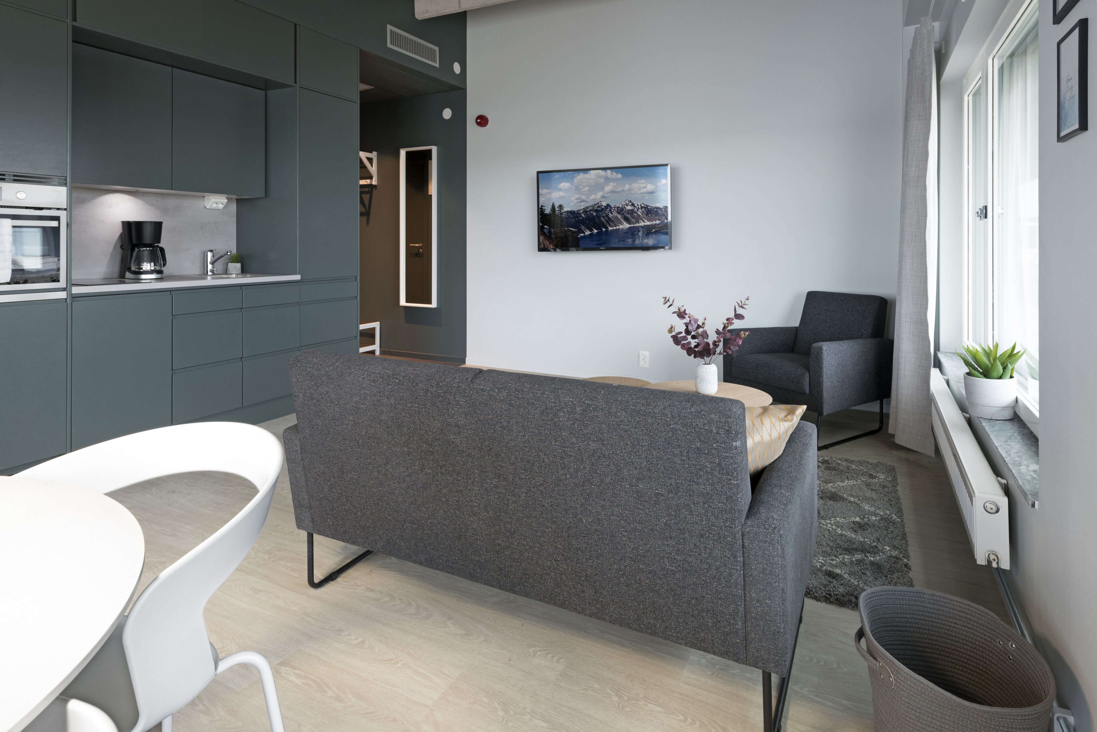 A modern living room with a grey sofa, a white dining table, and a kitchen area with grey cabinets and a coffee machine.