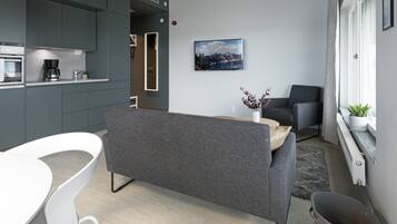 Un salon moderne avec un canapé gris, une table à manger blanche et un coin cuisine doté de meubles gris et d’une machine à café.