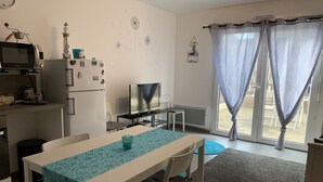 A compact living space with a kitchenette, a dining area with a table and chairs, a television, and a large window with curtains. - Charming Cottage Near Quend Beach: 2BR, Garden, Parking (Quend)