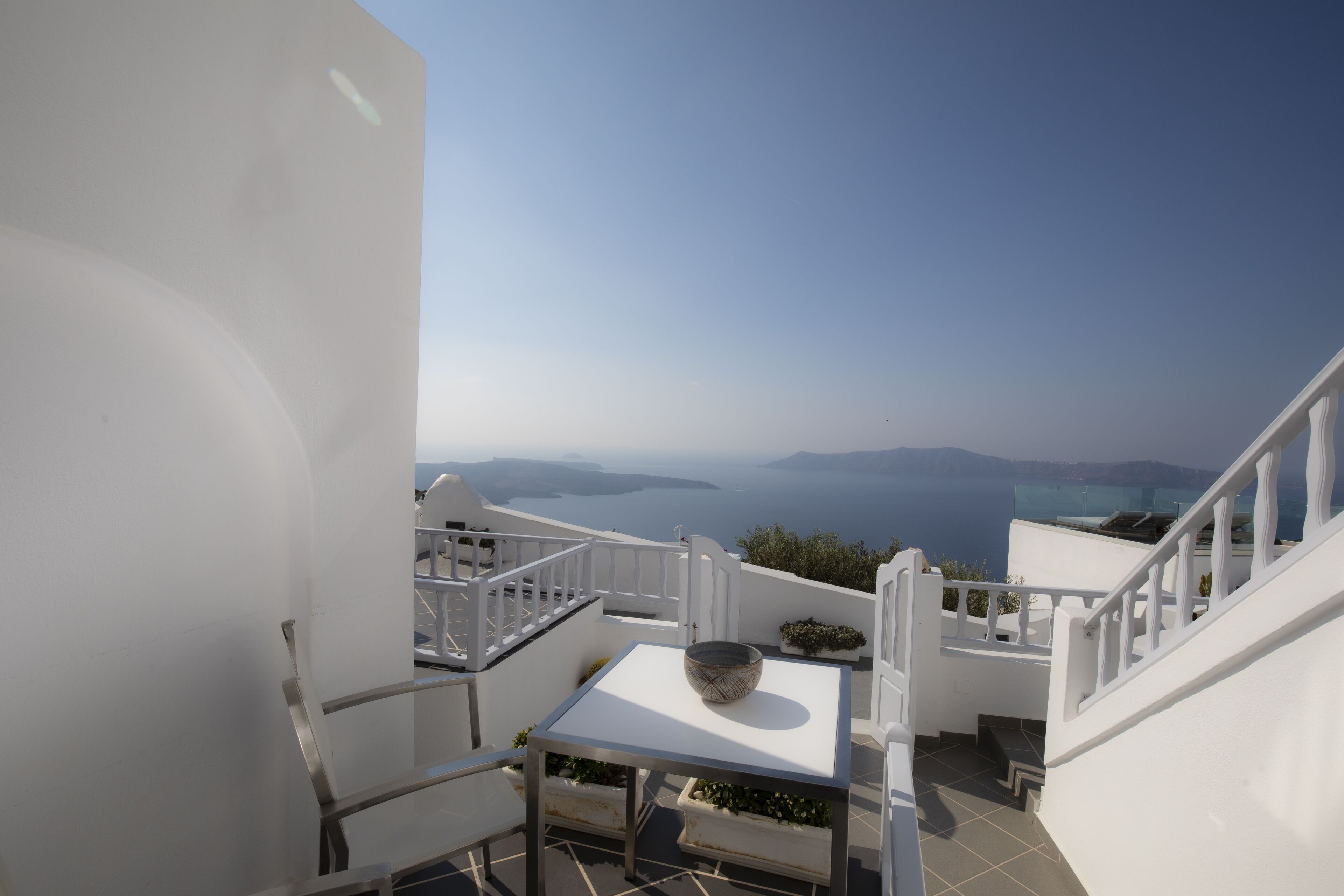 Una terrazza esterna bianca con tavolo e sedie, affacciata su una vista panoramica del mare e isole in lontananza.