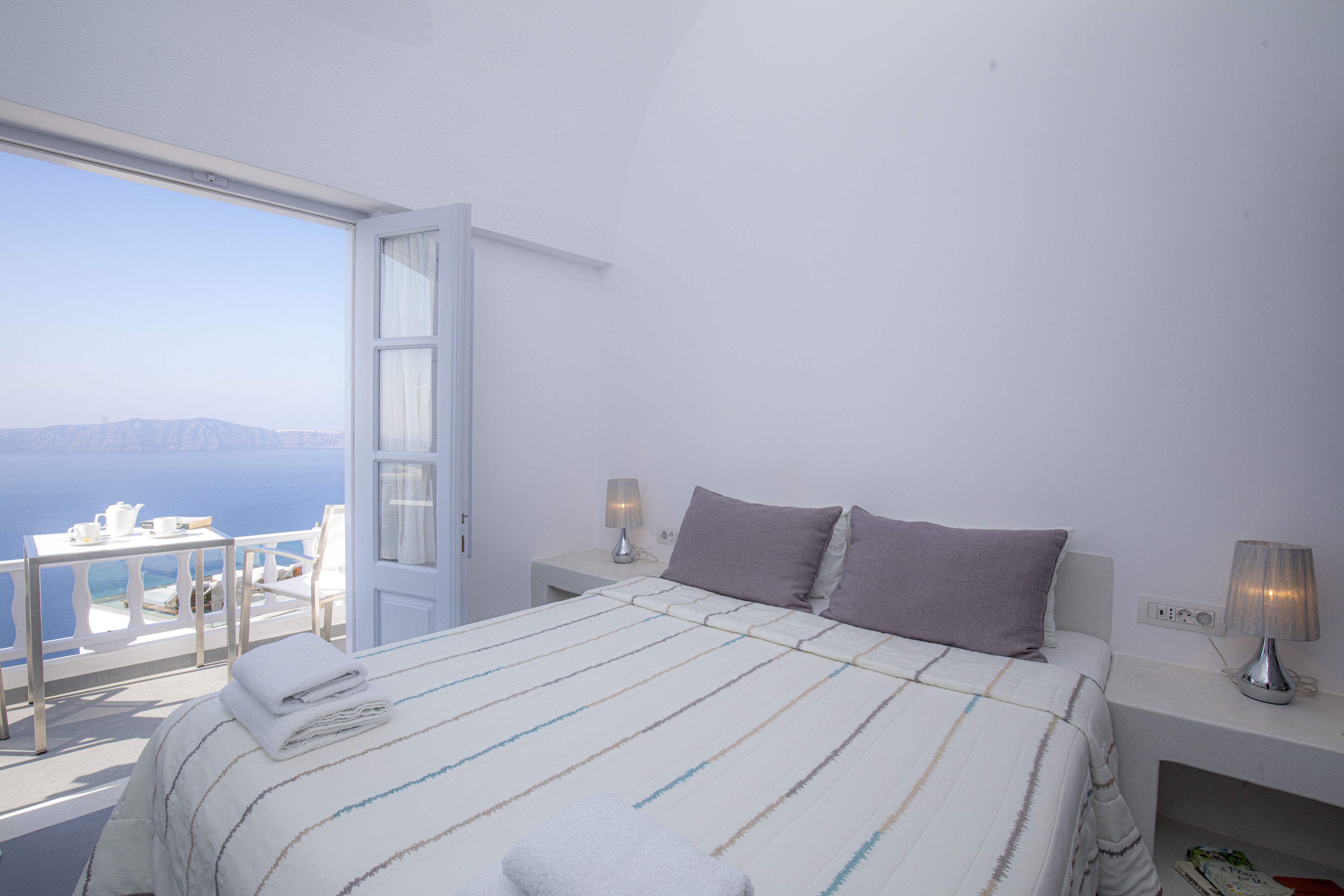 A bedroom with a bed, pillows, bedside tables, and a view of the sea.