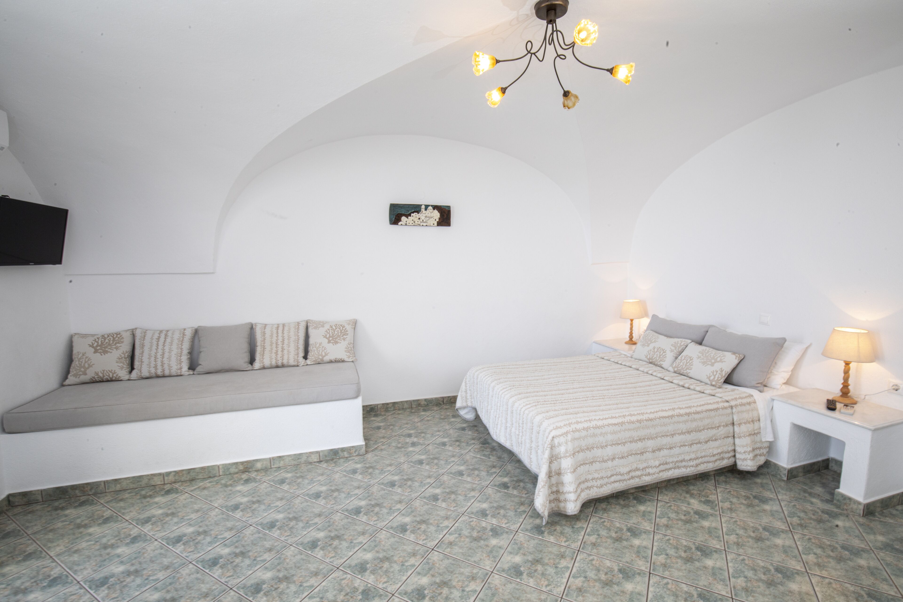 A bedroom with a bed, bedside tables, a bench, a chandelier, and a wall-mounted TV.