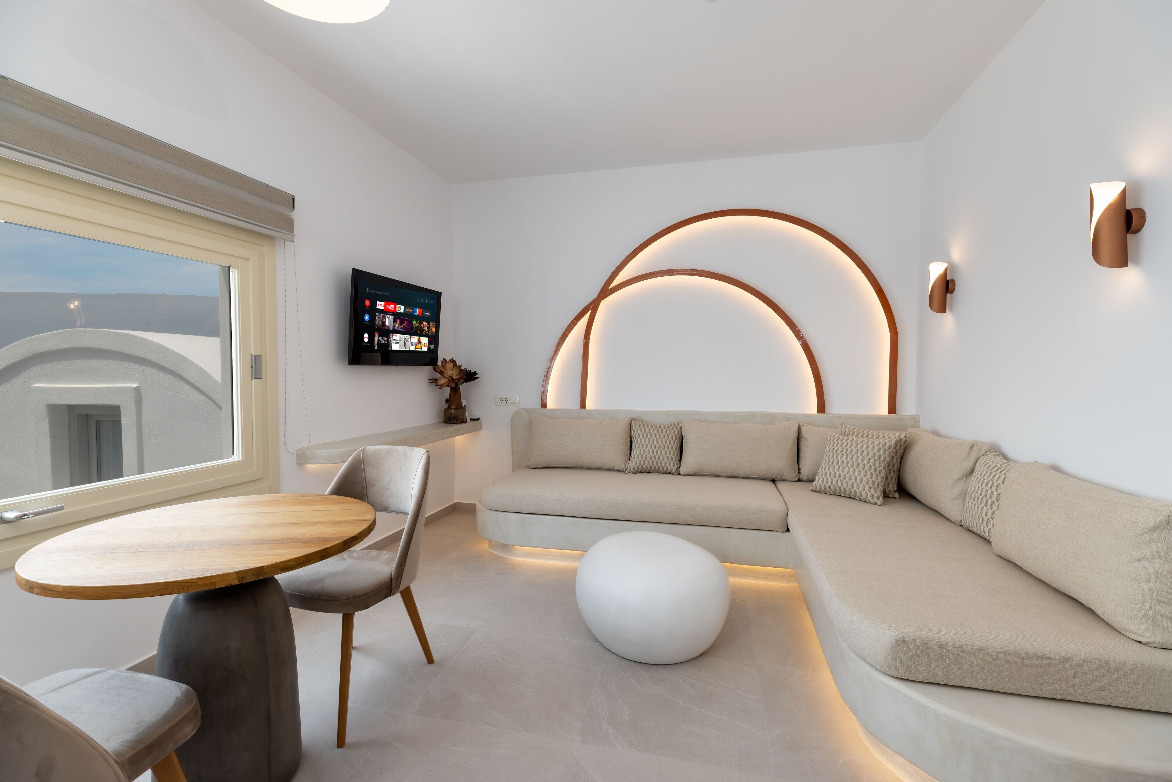 Una sala de estar moderna con sofá, mesa de centro redonda y sillones. Un televisor está montado en la pared y hay una ventana con vista a la ciudad.
