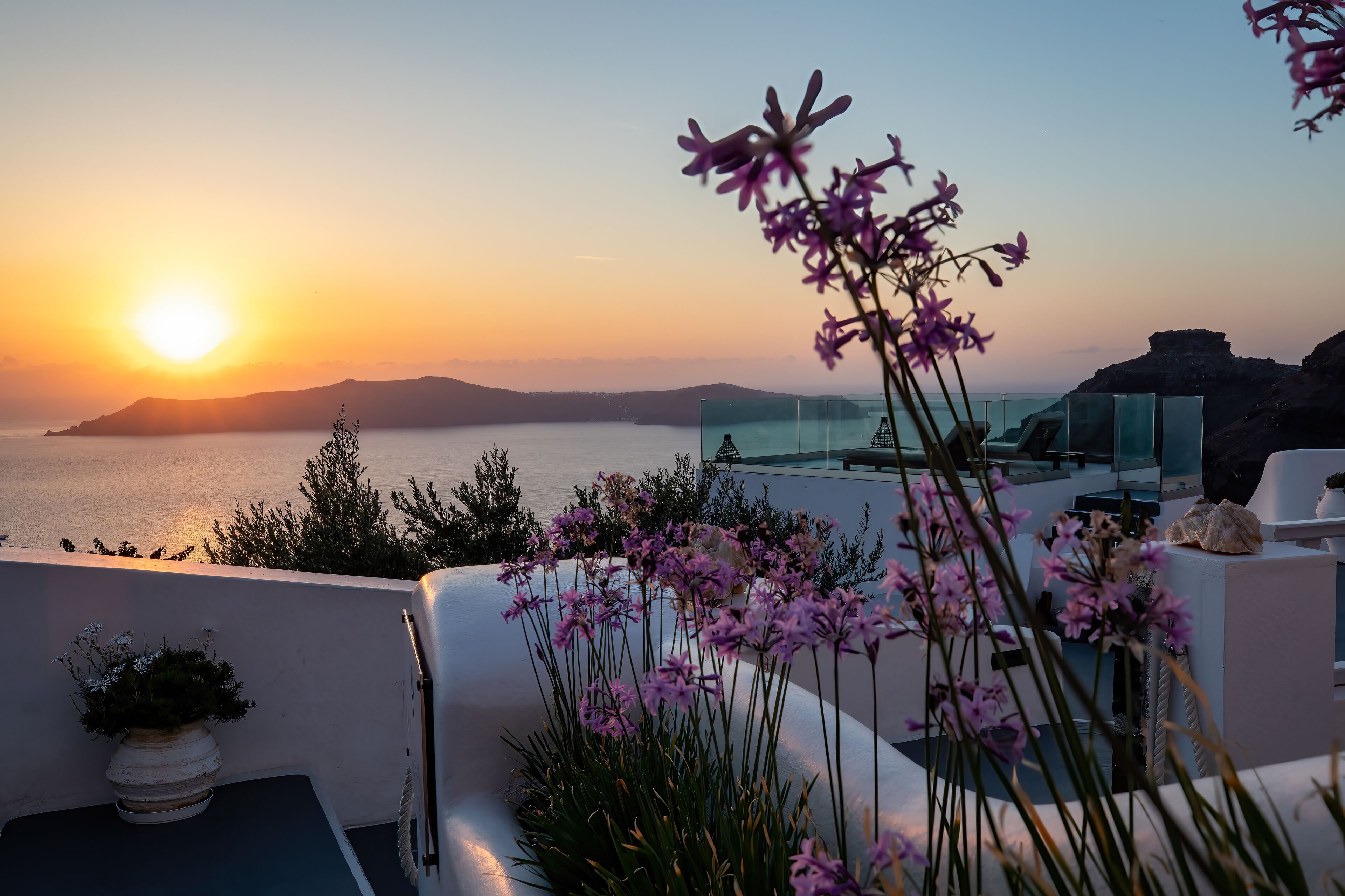 A modern rooftop terrace with a view of the sea and sunset, featuring potted plants and minimalist architecture.