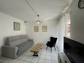 A living room with a grey sofa, a wooden bench, a black chair, a hanging light fixture, and two framed pictures on the wall.