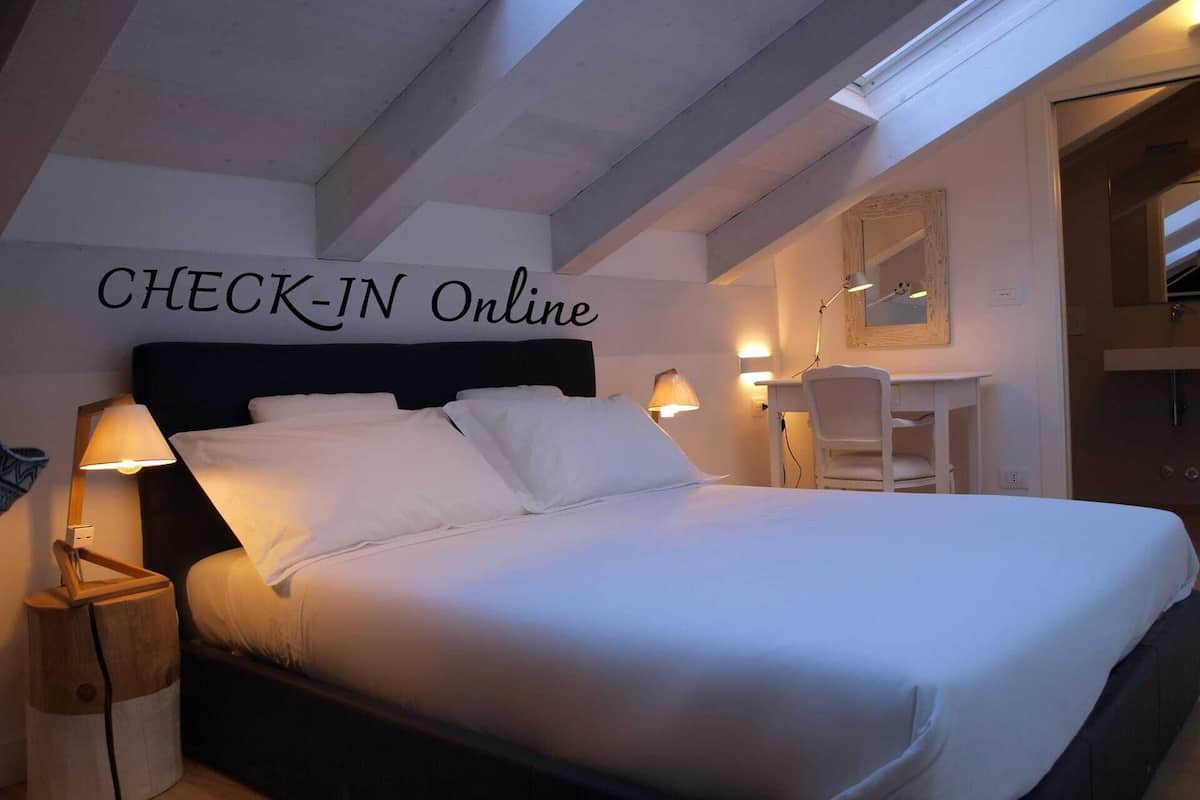 A hotel room with a bed, bedside lamps, a desk, and a mirror. The wall has a sign that says "CHECK-IN Online".