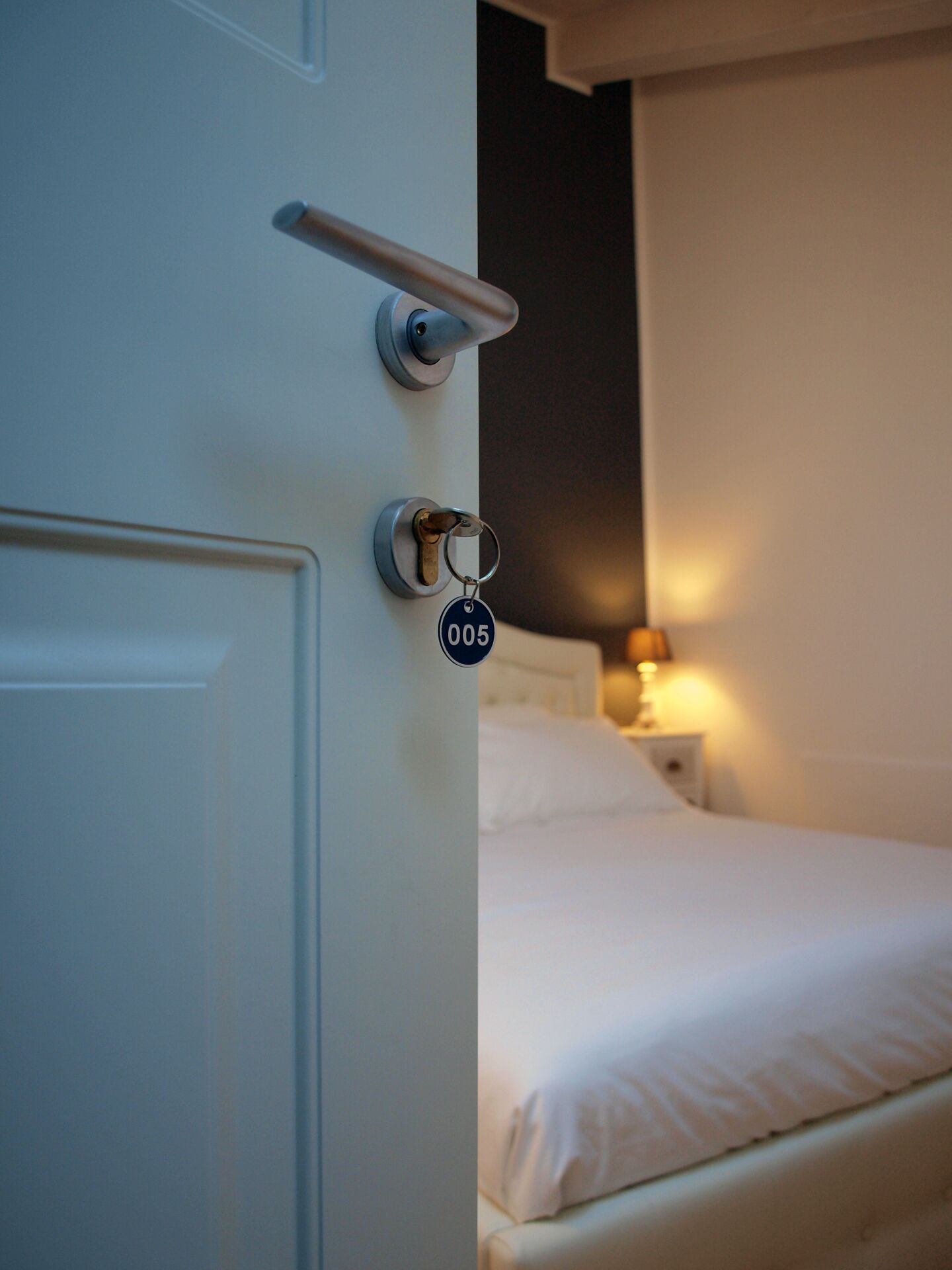A hotel room with a bed, a bedside table with a lamp, and a door with a keychain.