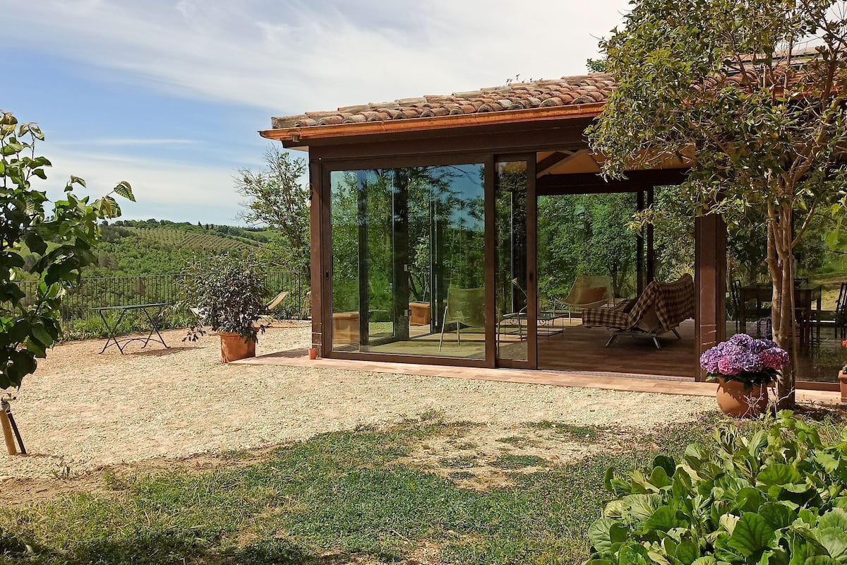 A glass-enclosed patio with a pergola, surrounded by a garden with potted plants and a view of rolling hills.