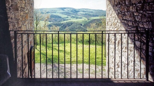 Un balcon aux murs de pierre, doté d’une balustrade en métal, qui offre une vue sur une vallée verdoyante.