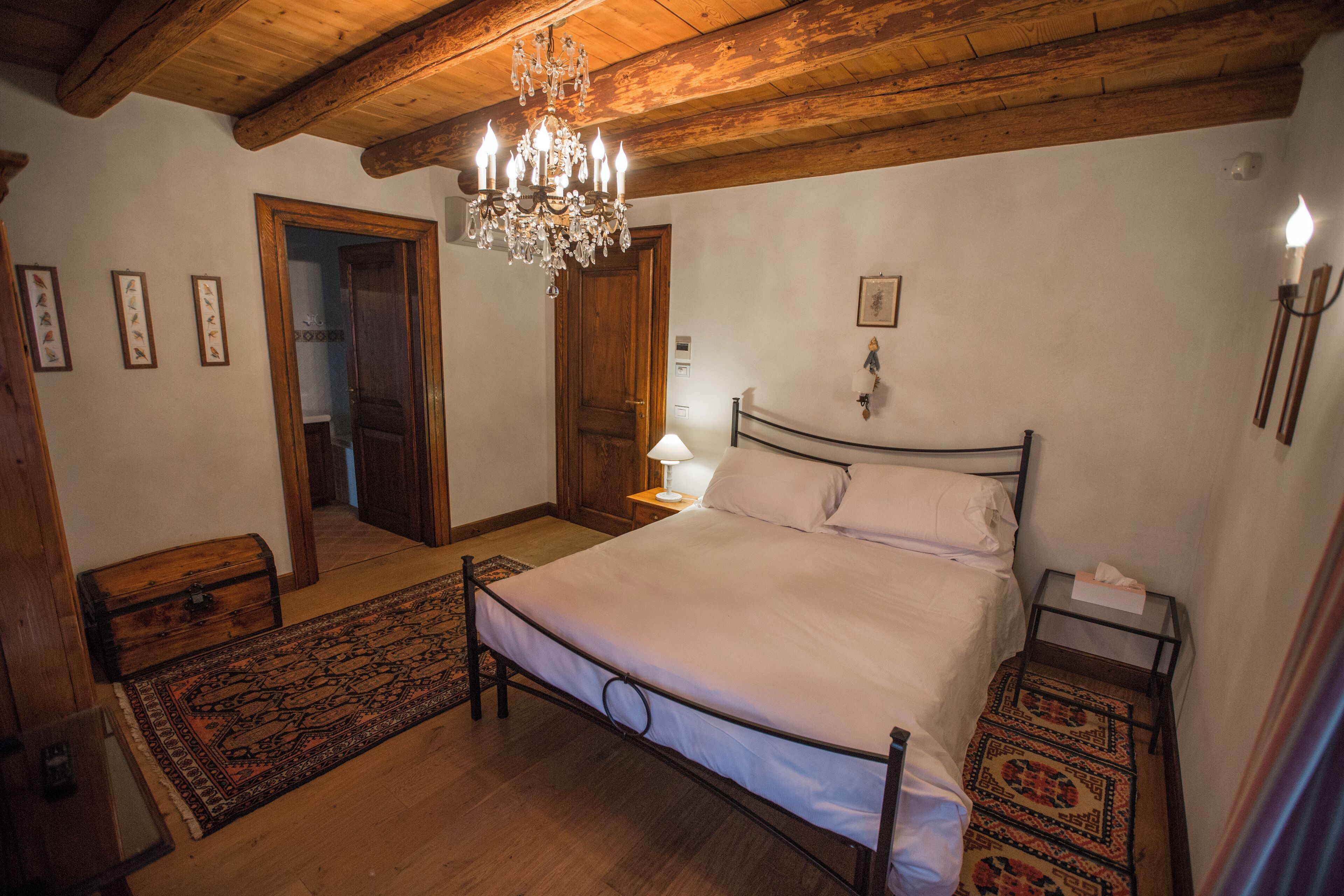 Un dormitorio con techo de madera, un candelabro, una cama con sábanas blancas, una mesita de noche y una alfombra estampada.