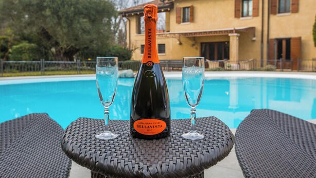 A bottle of Bellavista Brut champagne on a table with two glasses, by a pool with a house in the background.