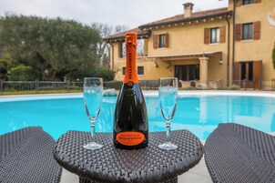 Una botella de champán Brut Bellavista sobre una mesa con dos copas, junto a una piscina con una casa al fondo.