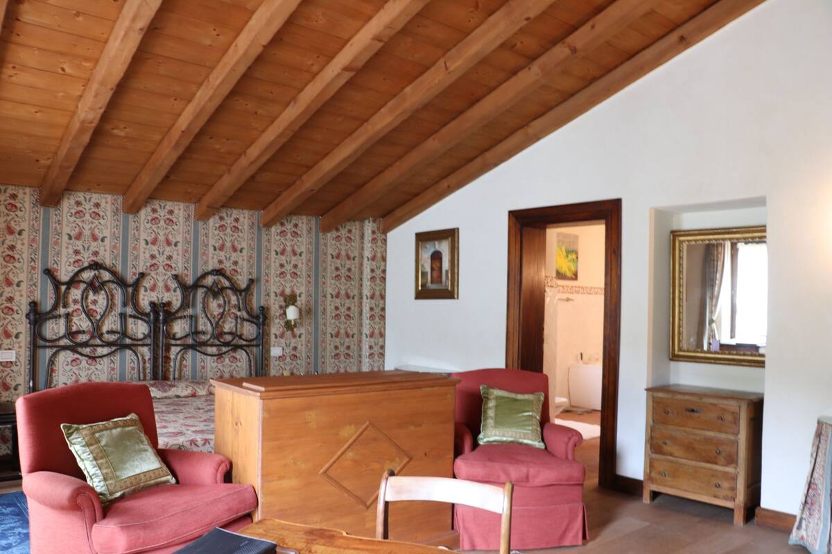 A room with wooden beams, floral wallpaper, a bed with a metal headboard, two red armchairs, a wooden dresser, and a mirror on the wall.