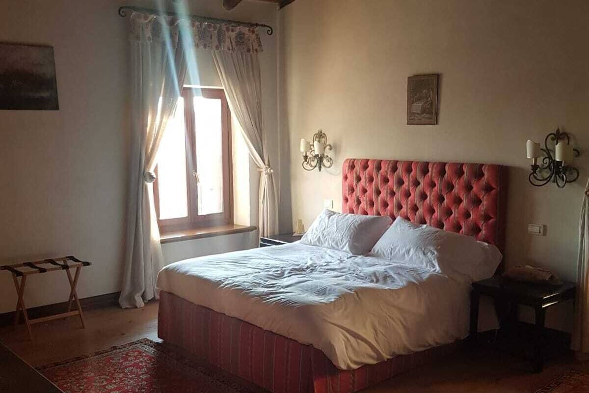 A bedroom with a wooden ceiling, a large bed with a red headboard, a nightstand, a small table, and a window with curtains.