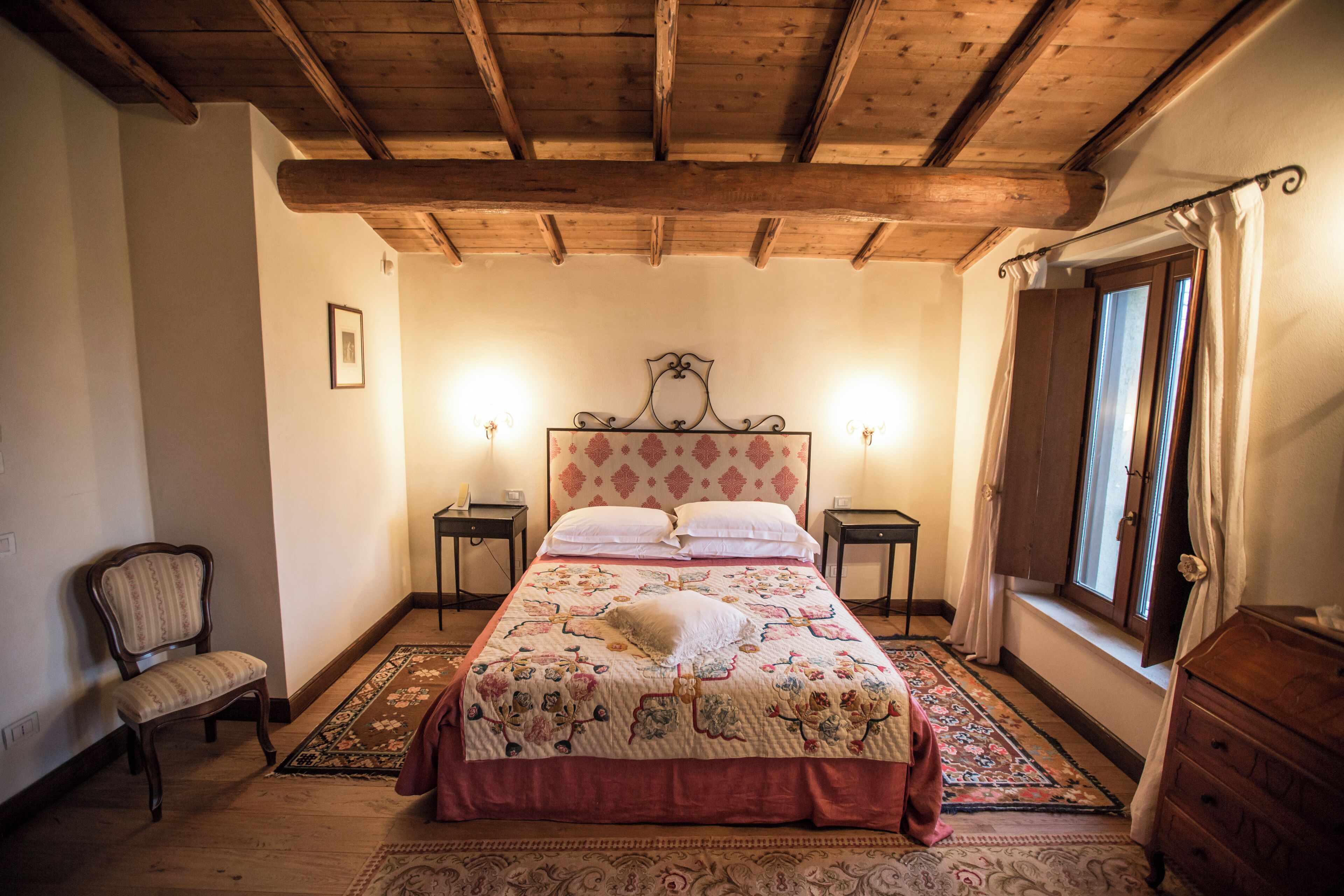 A bedroom with a wooden ceiling, a bed with a floral bedspread, two bedside tables with lamps, a chair, and a window with curtains.