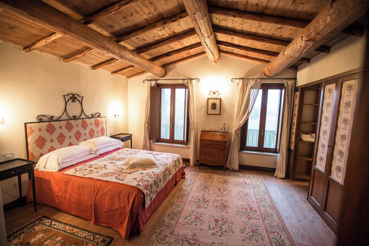A bedroom with a wooden ceiling, a bed with a floral bedspread, a nightstand, a dresser, and a window with curtains.