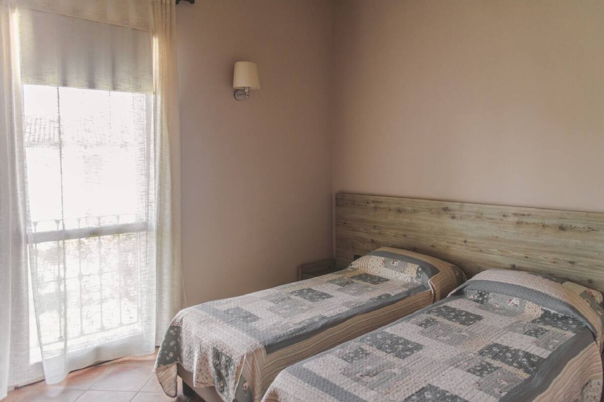 A room with two beds, a window with curtains, and a wall-mounted lamp.