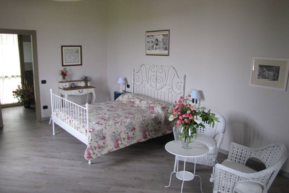 A bedroom with a bed, a bedside table, a chair, a small table, and a vase with flowers.