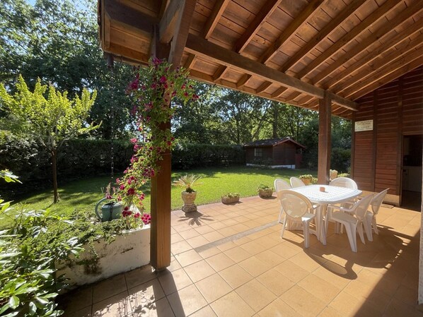 Une terrasse couverte avec une table et des chaises, entourée de verdure et avec un petit bâtiment en arrière-plan.