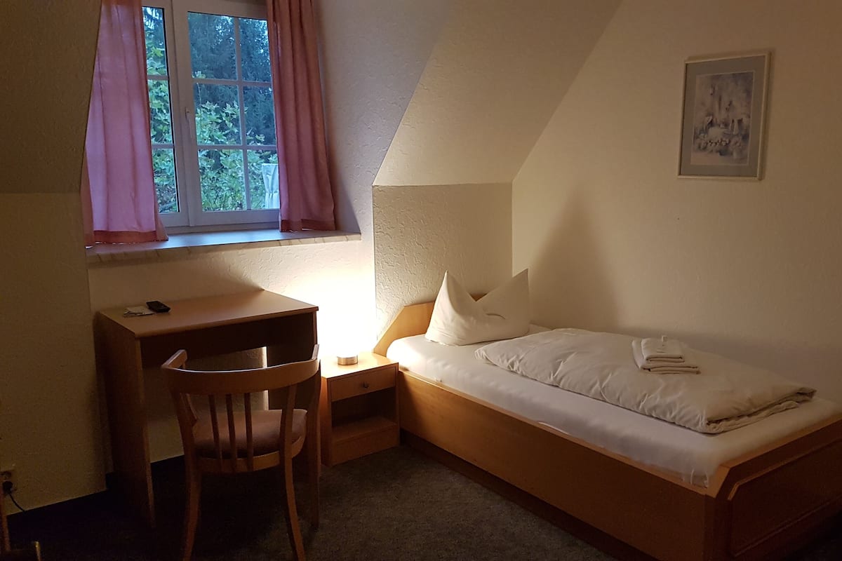 Una habitación pequeña con cama individual, escritorio y silla, una ventana con cortinas y un cuadro en la pared.