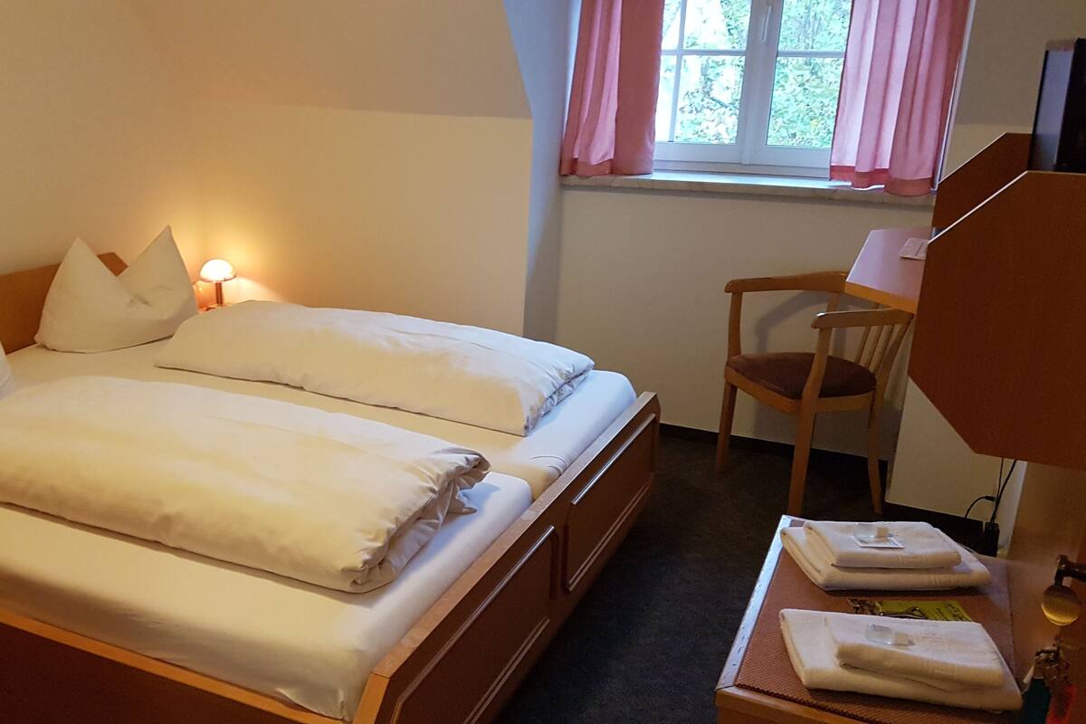 Una cama doble con sábanas blancas, una mesita de noche con lámpara, una silla y una ventana con cortinas.