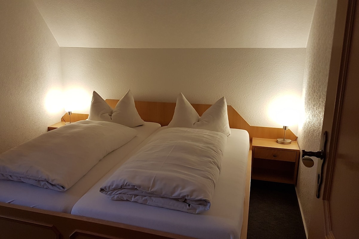 Una cama doble con ropa de cama y almohadas blancas, dos mesitas de noche con lámparas y un cabecero de madera.
