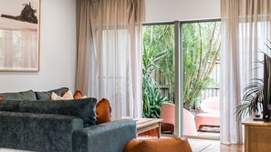 Ein Wohnzimmer mit einer Couch, einem Couchtisch und einer TV-Kommode. Eine Glastür öffnet sich zu einem Außenbereich mit Pflanzen.