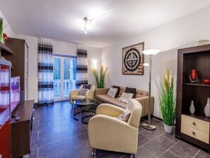 A modern living room with a sofa, armchairs, a coffee table, and a floor lamp. There is a large window with curtains, a plant, and a wall-mounted artwork. - (204) 2-room apartment - Apartment house Villa Laura (Kühlungsborn)
