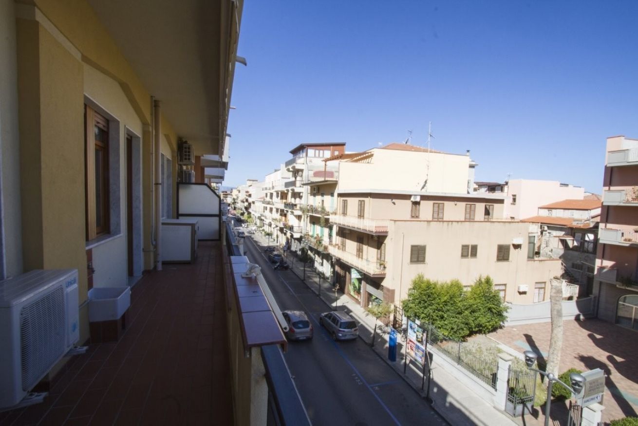 Ein schmaler Balkon mit Blick auf eine Straße mit mehreren Gebäuden. Der Balkon hat einen gefliesten Boden und ein weißes Geländer.